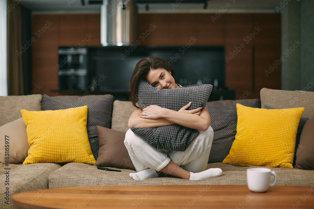 Obraz premium Happy young girl hugging soft pillow sitting on comfortable sofa. Relaxed female resting in modern living room at home