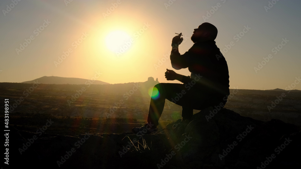 contour or silhouette of man against background of sunset in mountains, man sits on edge of mountain and lights cigarette, enjoys view.