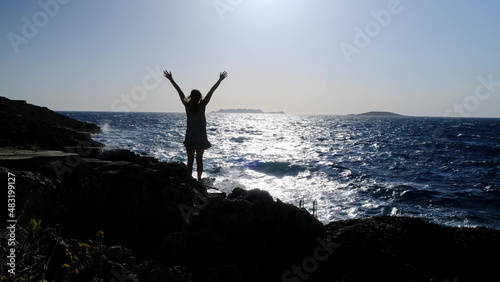 slender blonde in striped dress stands against backdrop of sea or ocean and looks into distance. woman in dress stands on rocky shore of reservoir in windy weather, hands up, wave beat
