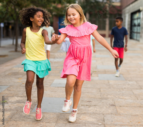 Happy blonde preteen girl enjoying walk with african american girl friend along city street on summer day, running skipping gaily and holding hands. Childhood friendship concept