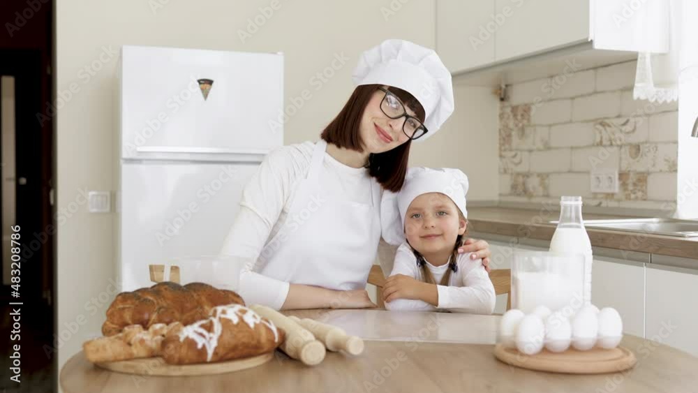 Adorable Little Girl And Her Caucasian Mom Cooking In Kitchen. Happy mom and her Female Kid hugging and looking at the camera and preparing for Kneading Dough For Cookies Together, having Fun At Home Adorable Little Girl And Her Caucasian Mom Cooking In Kitchen. Happy mom and her Female Kid hugging and looking at the camera and preparing for Kneading Dough For Cookies Together, having Fun At Home