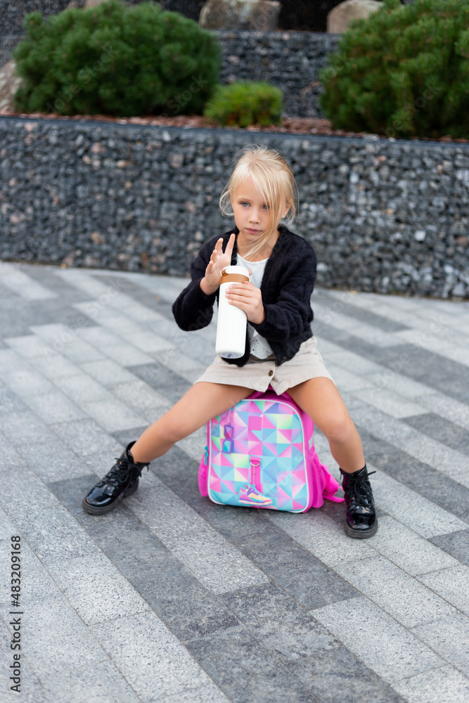 Happy blonde girl sitting on backpack drinking tea from thermos cup.  Schoolgirl girl drinks water from a thermos or thermomug.