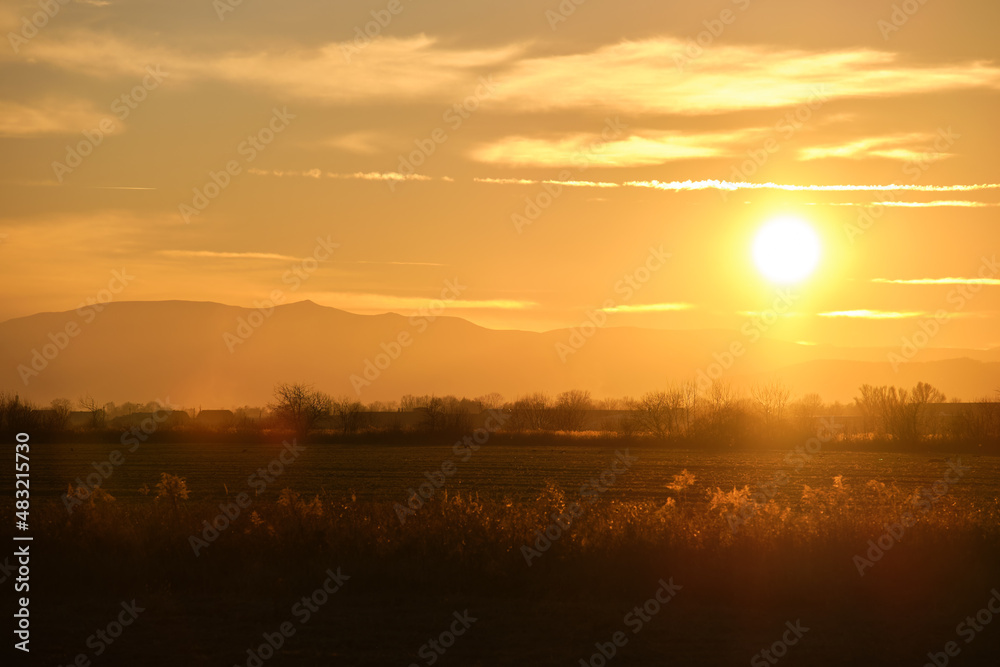 Beautiful evening panoramic landscape with bright setting sun over distant mountain peaks and asphalt road in front at sunset Beautiful evening panoramic landscape with bright setting sun over distant mountain peaks and asphalt road in front at sunset