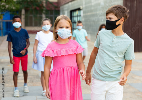 European boy and girl in face masks walking together hand in hand through city streets in summertime. Other kids walking behind.