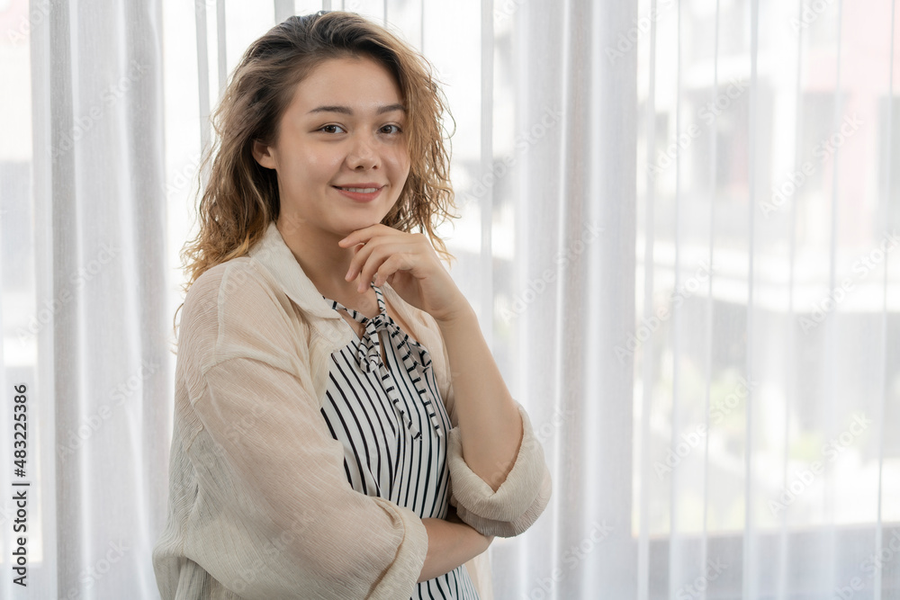Young woman stand by the window with smiling on her face with the pose of folded right arm and left arm under her chin, lifestyle confidence concept