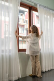 The back shot of young woman open the white curtain for the morning sunshine. She stands by the window with the pose of  open white sheer curtain with her arms. Lifestyle optimism concept