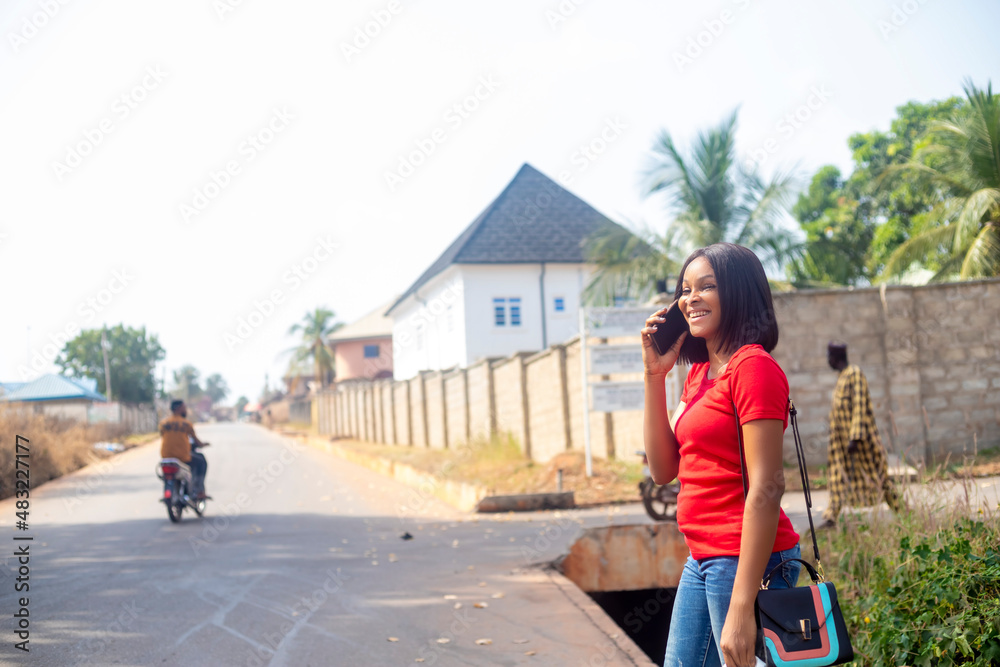 Young black african woman holding mobile phone, using smartphone, making a call, talking on the phone, standing on sunny street waiting for cab taxi