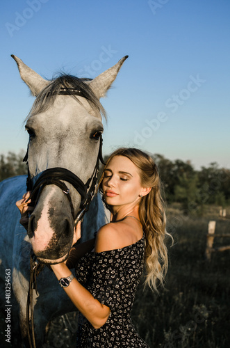 Portrait of a beautiful young blonde woman with closed eyes near a white horse in the summer on the field close-up. Vertical