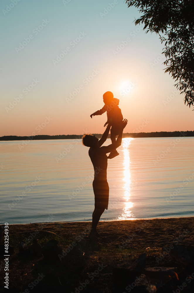 Fatherhood, childhood, family vacation. Silhouettes of father and son having fun together on beach at sunset, outdoors. Side view of dad holding child in arms, throwing up air against sky