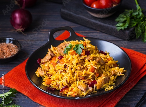 Pilaf, a dish of rice, chicken, carrots with spices and dried cranberries on a black plate on a dark wooden background.