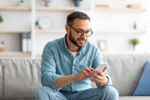 Positive young guy in eyeglasses sitting on sofa with mobile phone and texting to his friend, indoors