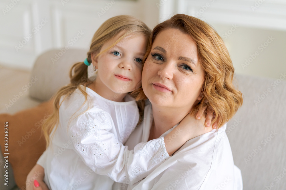 European mom and little daughter are hugging while sitting on sofa in bright living room at home. The relationship between children and parents. Happy motherhood European mom and little daughter are hugging while sitting on sofa in bright living room at home. The relationship between children and parents. Happy motherhood
