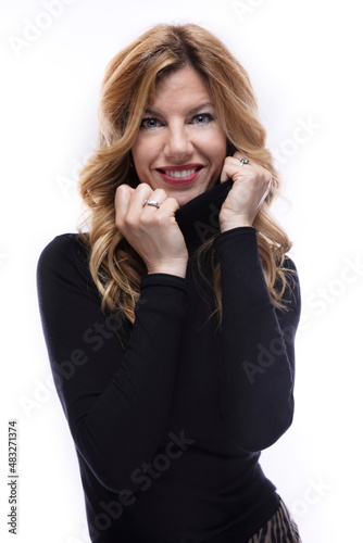 Blonde woman dressed in a black turtleneck sweater and looking directly at the camera, while smiling and partially covering herself with her sweater, on white background.