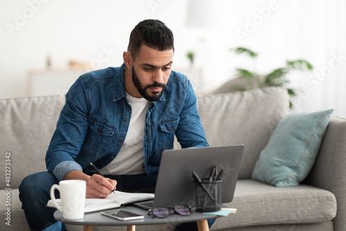Serious young Arab man using laptop, working remotely, taking notes during remote business meeting from home