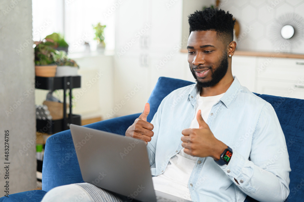 Cheerful multiracial guy taking a part in virtual meeting sitting with a laptop on laps on sofa at home, talking at the webcam and showing thumbs up. Male student during online classes