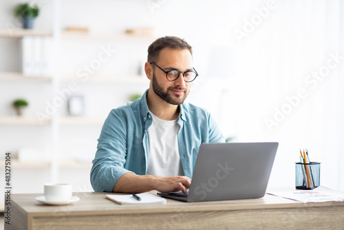 Young Caucasian man in eyeglasses working online, sitting at desk and using laptop from home office