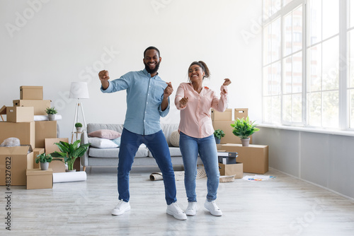 Satisfied young afro american couple dancing in flat with cardboard boxes in home interior, free space