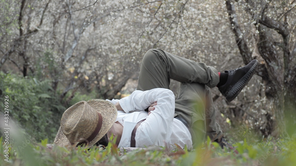 Man relaxing under a tree, laying down and resting using a hat to cover his face, man in white with pose lying on grass rest concept