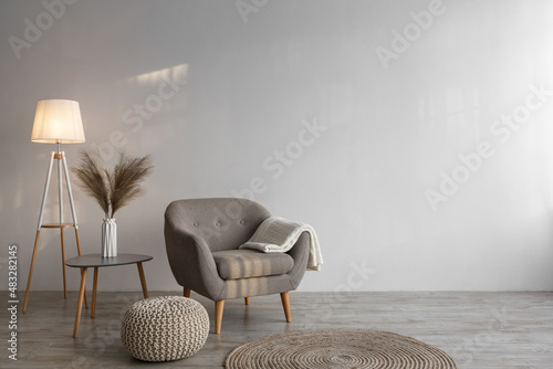 Fotografie, Obraz Retro armchair with plaid, luminous lamp, dry plants in vase on table, ottoman a