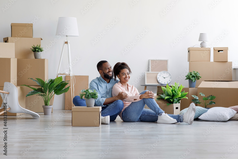Cheerful young black husband and wife rest, sit on floor among cardboard boxes with things and plants Cheerful young black husband and wife rest, sit on floor among cardboard boxes with things and plants