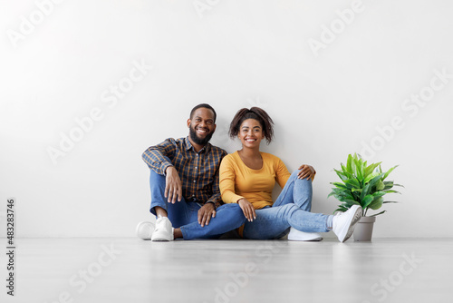 Glad young african american man and lady sit on floor with plant in pot on white wall background, rest after relocation