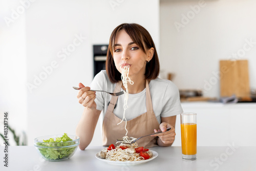 Happy lady eating spaghetti and vegetable salad, enjoying tasty lunch, sitting at table and looking at camera in kitchen