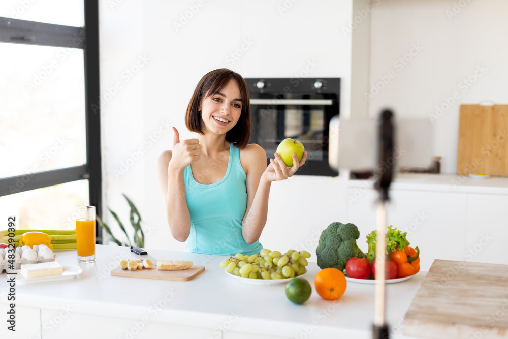 Happy woman showing thumb up and holding apple, recording new video for sport and food blog, kitchen interior Happy woman showing thumb up and holding apple, recording new video for sport and food blog, kitchen interior