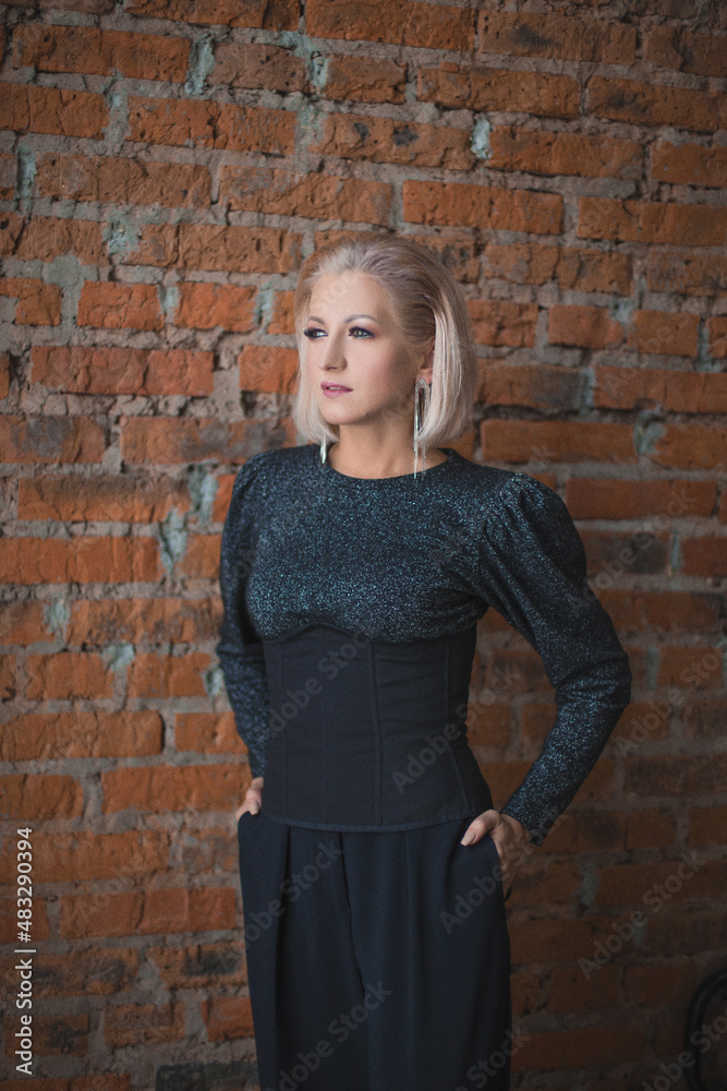 blonde girl in a shiny sweater, black corset and black trousers stands near a brick wall