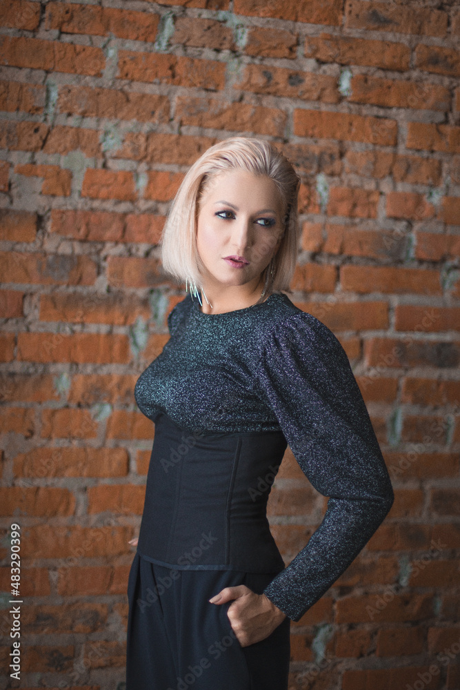 blonde girl in a shiny sweater, black corset and black trousers stands near a brick wall