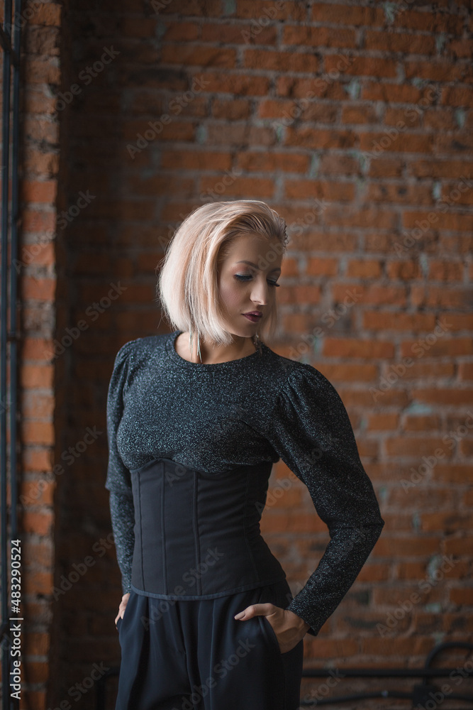 blonde girl in a shiny sweater, black corset and black trousers stands near a brick wall