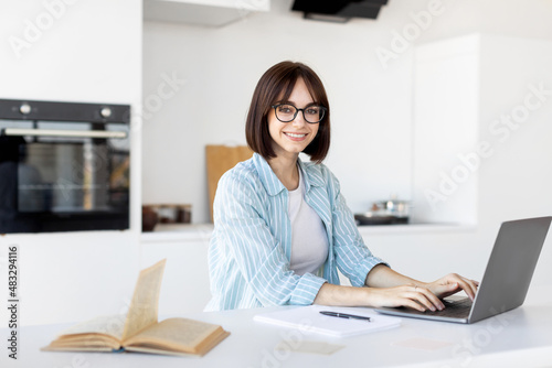 Happy young freelancer working on laptop in kitchen interior, sitting at table and smiling at camera