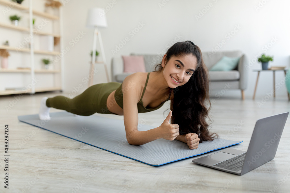 Obraz premium Online sports. Millennial Indian woman standing in elbow plank in front of laptop, showing thumb up gesture at home