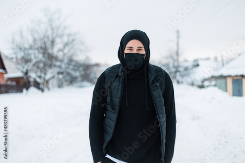 handsome young stylish hipster man with a protective mask in black fashionable outerwear with a hoodie, vest, hat and backpack walks on a winter day with snow. Guy in the town. Coronavirus concept