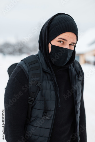 Handsome young man with protective medical mask in black outerwear with vest, black hoodie, hat and backpack travels on a winter day with snow