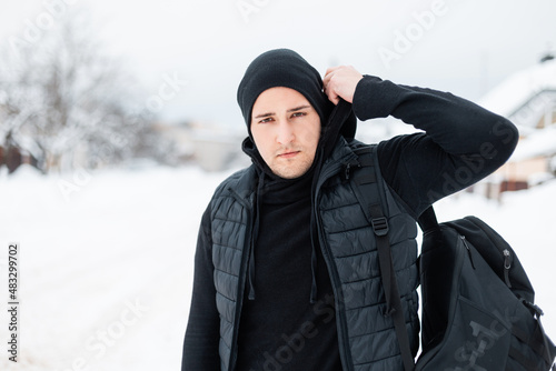 Handsome young stylish man in black fashionable dress with a vest, a hoodie, a hat and a backpack walks on the street on a winter snowy day
