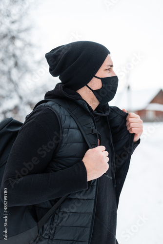 Handsome stylish hipster guy with a black textile protective mask in fashionable winter clothes with a backpack, vest and hat walks on the street with snow. Male style and pandemic fashion concept