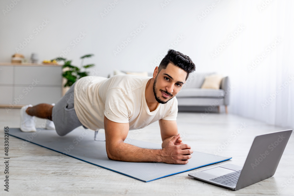 Fototapeta premium Online sports during covid lockdown. Strong young Arab guy doing exercises in front of laptop at home