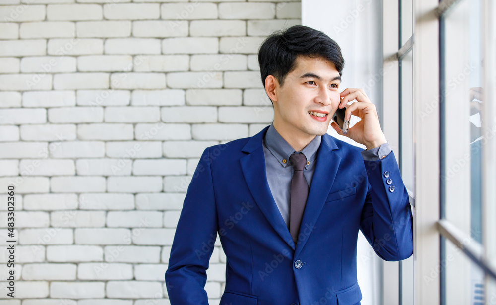 Portrait studio shot of Asian handsome professional successful male businessman entrepreneur in formal blue suit necktie standing holding smartphone smiling on white brick wall background. Portrait studio shot of Asian handsome professional successful male businessman entrepreneur in formal blue suit necktie standing holding smartphone smiling on white brick wall background.