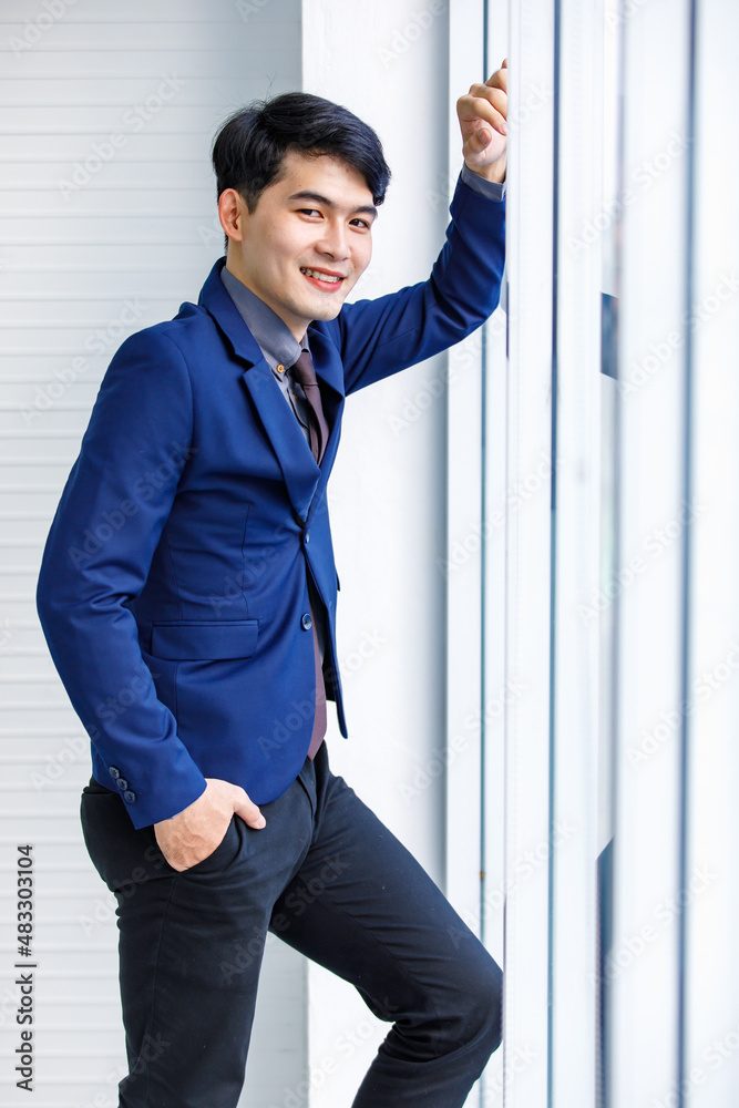Millennial Asian handsome smart confident professional success male manager businessman entrepreneur in formal blue suit smiling standing near skyscraper office window