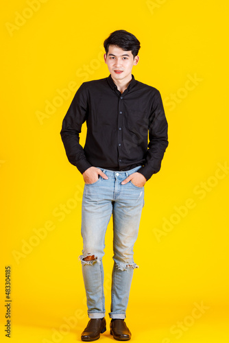 Portrait studio shot of millennial Asian professional successful male businessman in casual black long sleeve shirt outfit standing holding hand in tearing denim jeans pocket on yellow background