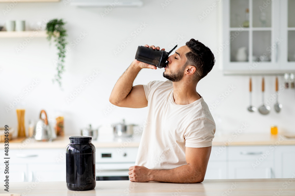 Young Arab guy drinking protein shake from bottle at kitchen, copy space. Body care concept - obrazy, fototapety, plakaty 