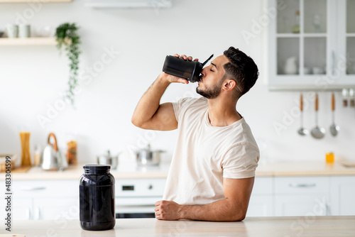 Young Arab guy drinking protein shake from bottle at kitchen, copy space. Body care concept photo