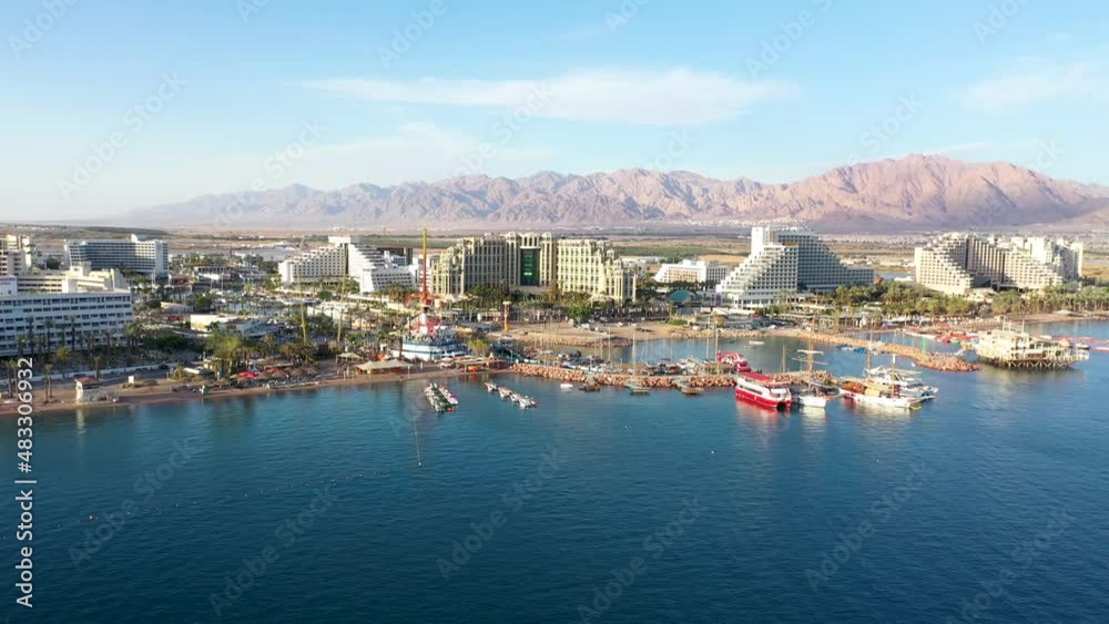 Drone view of Eilat Israel on sunny clear day from the sea flying in towards boats, luxury hotels and resort area with beach port marina and promenade. Desert and mountains are in the background. Drone view of Eilat Israel on sunny clear day from the sea flying in towards boats, luxury hotels and resort area with beach port marina and promenade. Desert and mountains are in the background.