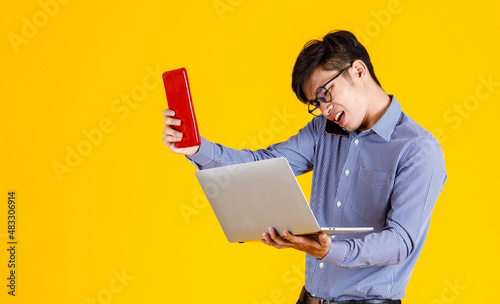 Studio shot of millennial busy stressed overload Asian professional male businessman employee holding laptop computer and tablet in hands on call with customer via smartphone on yellow background