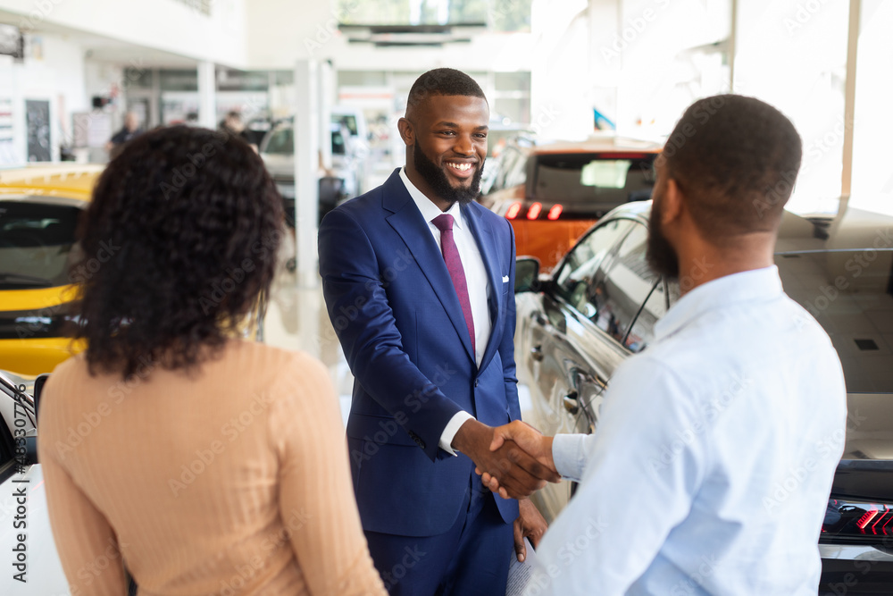 Car Dealership Concept. Salesperson Handshaking With Young Black Customers Couple In Showroom Car Dealership Concept. Salesperson Handshaking With Young Black Customers Couple In Showroom