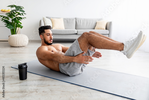 Shirtless young Arab man working out core muscles, doing abs exercises on yoga mat at home, full length photo