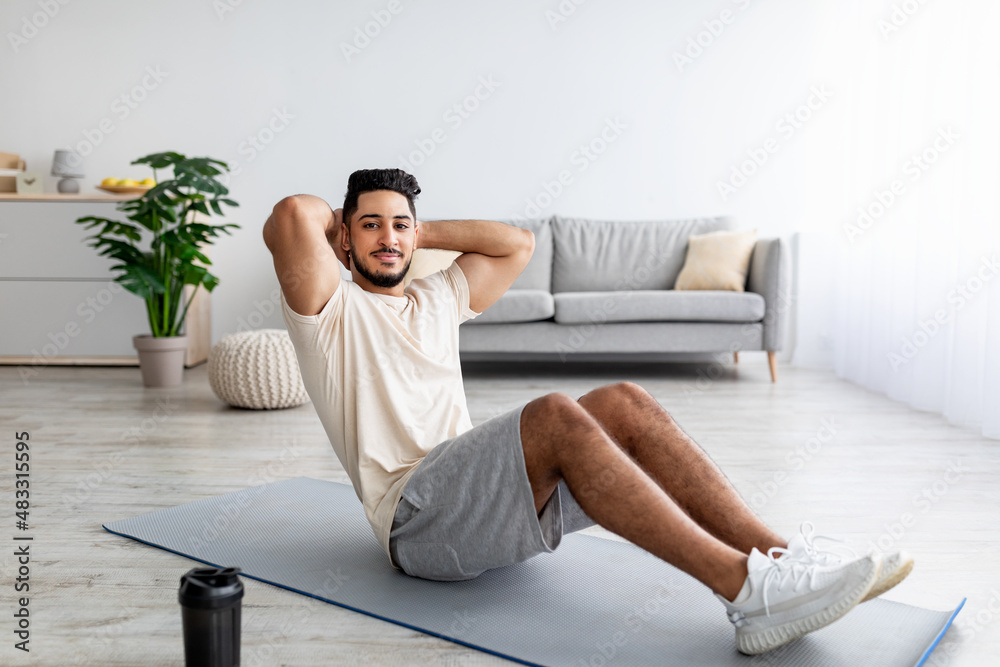Athletic millennial Arab man doing abs exercises, working out at home, full length