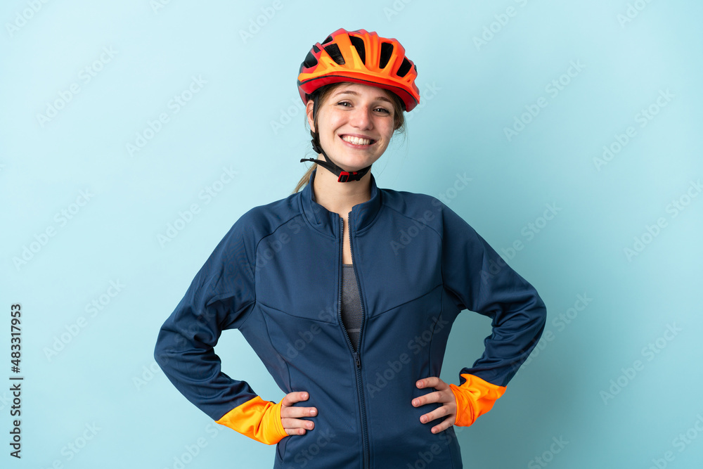 Young cyclist woman isolated on blue background posing with arms at hip and smiling - obrazy, fototapety, plakaty 
