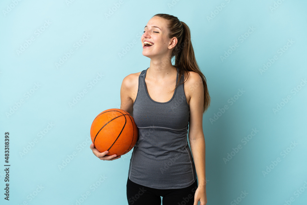 Young woman playing basketball isolated on blue background laughing in lateral position