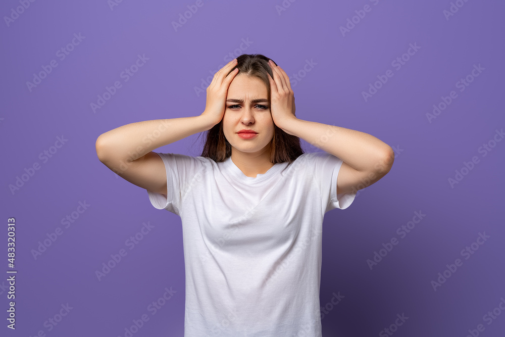 Shocked concerned young woman, hold hands on head, look worried and concerned as facing difficulties, standing in blank white t shirt over purple background Shocked concerned young woman, hold hands on head, look worried and concerned as facing difficulties, standing in blank white t shirt over purple background